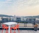 A rooftop setting features a modern white table accompanied by two vibrant orange chairs, overlooking a cityscape in the distance. The background consists of a variety of buildings and a skyline, with hills and a bridge. There is a potted plant on the right side, and the sky is lightly cloudy with a hint of sunset.