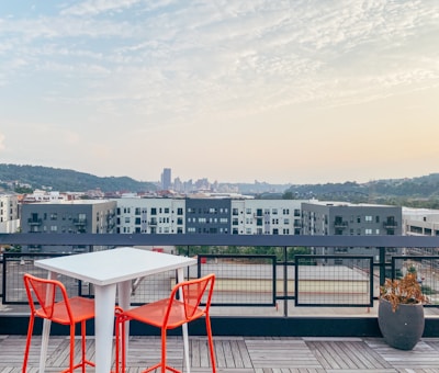 A rooftop setting features a modern white table accompanied by two vibrant orange chairs, overlooking a cityscape in the distance. The background consists of a variety of buildings and a skyline, with hills and a bridge. There is a potted plant on the right side, and the sky is lightly cloudy with a hint of sunset.