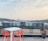 A rooftop setting features a modern white table accompanied by two vibrant orange chairs, overlooking a cityscape in the distance. The background consists of a variety of buildings and a skyline, with hills and a bridge. There is a potted plant on the right side, and the sky is lightly cloudy with a hint of sunset.