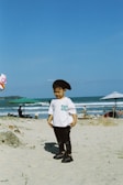 A young child stands on a sandy beach wearing a white t-shirt with the word 'SICK' in green letters, black pants, and black shoes. The child is also wearing a black beret and appears to be looking downwards. The background features the ocean with waves, and several beach umbrellas are visible. There are a few people in the distance enjoying the beach.