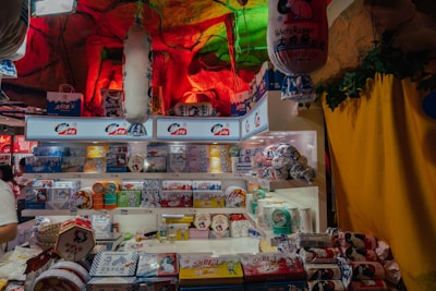 A store display featuring a variety of White Rabbit branded products. The shelves are filled with colorful packaging displaying images of rabbits and logos. The background has a cave-like appearance with red and green lighting, giving an atmospheric feel. Decorative elements like hanging lanterns and leafy accents add to the visual interest.