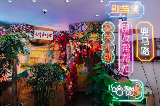 The image features a vibrant interior space adorned with colorful decorations, including hanging flowers and leaves in shades of red, purple, and pink. Bright neon signs with Chinese characters are prominently displayed, adding an urban and lively atmosphere. Lush green plants are placed around the area, contributing to a tropical and inviting ambiance.