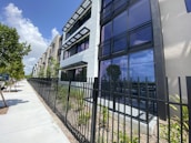 A modern apartment building with multiple stories, featuring large glass windows and balconies with black railing. The exterior is predominantly shades of white and gray. A black metal fence borders the property, with a sidewalk and small trees lining the street nearby. The sky is blue with some clouds, indicating a sunny day.