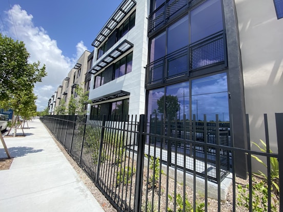 A modern apartment building with multiple stories, featuring large glass windows and balconies with black railing. The exterior is predominantly shades of white and gray. A black metal fence borders the property, with a sidewalk and small trees lining the street nearby. The sky is blue with some clouds, indicating a sunny day.