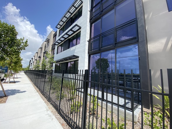 A modern apartment building with multiple stories, featuring large glass windows and balconies with black railing. The exterior is predominantly shades of white and gray. A black metal fence borders the property, with a sidewalk and small trees lining the street nearby. The sky is blue with some clouds, indicating a sunny day.