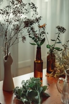 Various dried and fresh flowers are arranged in glass bottles and a ceramic vase shaped like a hand. The setting is on a wooden table near a softly lit window, creating a cozy and serene atmosphere.