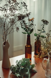 Various dried and fresh flowers are arranged in glass bottles and a ceramic vase shaped like a hand. The setting is on a wooden table near a softly lit window, creating a cozy and serene atmosphere.