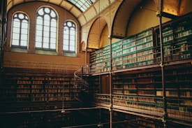 A grand library interior featuring tall bookshelves filled with a multitude of books, reaching up to arched windows. The ornate design of the ceiling and the intricate spiral staircase add to the atmosphere of elegance and grandeur.