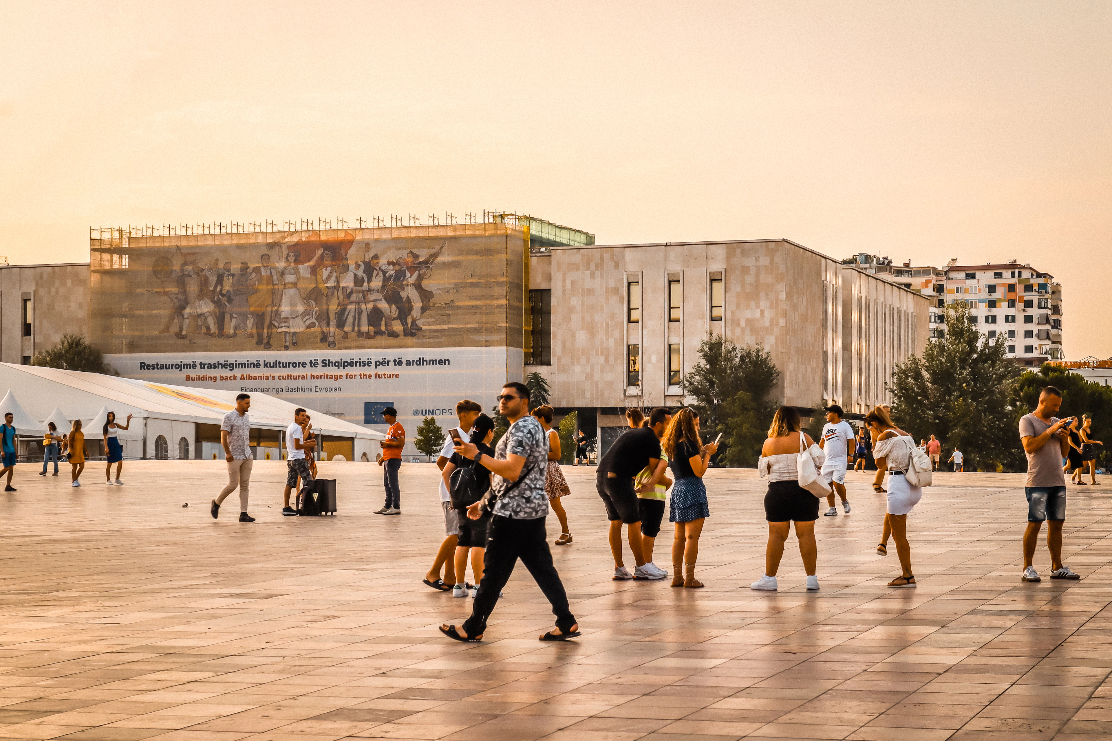People walk across Skanderbeg Square in Tirana at sunset, with the National History Museum mosaic in the background.