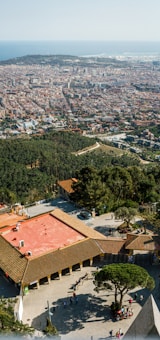 A panoramic view of a city by the sea, with dense urban structures spread across the landscape. A large green area with trees is visible in the foreground, along with a plaza containing a red-roofed building. A group of people can be seen gathering in the open area surrounded by the greenery and buildings.