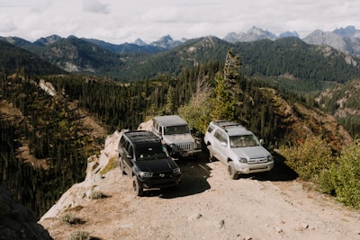 Three SUVs are parked on a dirt path at the edge of a cliff, surrounded by a picturesque mountain landscape. The forested hills and rocky peaks in the background create a scenic and adventurous atmosphere.