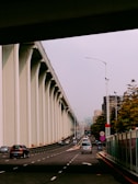 A wide road runs underneath an elevated highway with tall concrete columns to the left. Several vehicles, including cars and a bus, travel along the multi-lane road. High-rise buildings and trees are visible in the background, indicating an urban environment. Streetlights and traffic signs line the road.