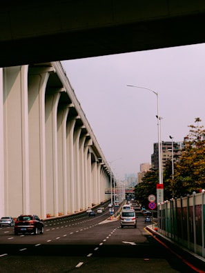A wide road runs underneath an elevated highway with tall concrete columns to the left. Several vehicles, including cars and a bus, travel along the multi-lane road. High-rise buildings and trees are visible in the background, indicating an urban environment. Streetlights and traffic signs line the road.