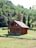 A small log cabin with a wooden exterior is situated in a lush, green landscape. The structure features a triangular roof, a front porch, and several windows. It is surrounded by dense forest with various shades of green, including tall trees and bushes. The foreground displays well-maintained grass, enhancing the serene and natural ambiance.