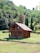 A small log cabin with a wooden exterior is situated in a lush, green landscape. The structure features a triangular roof, a front porch, and several windows. It is surrounded by dense forest with various shades of green, including tall trees and bushes. The foreground displays well-maintained grass, enhancing the serene and natural ambiance.