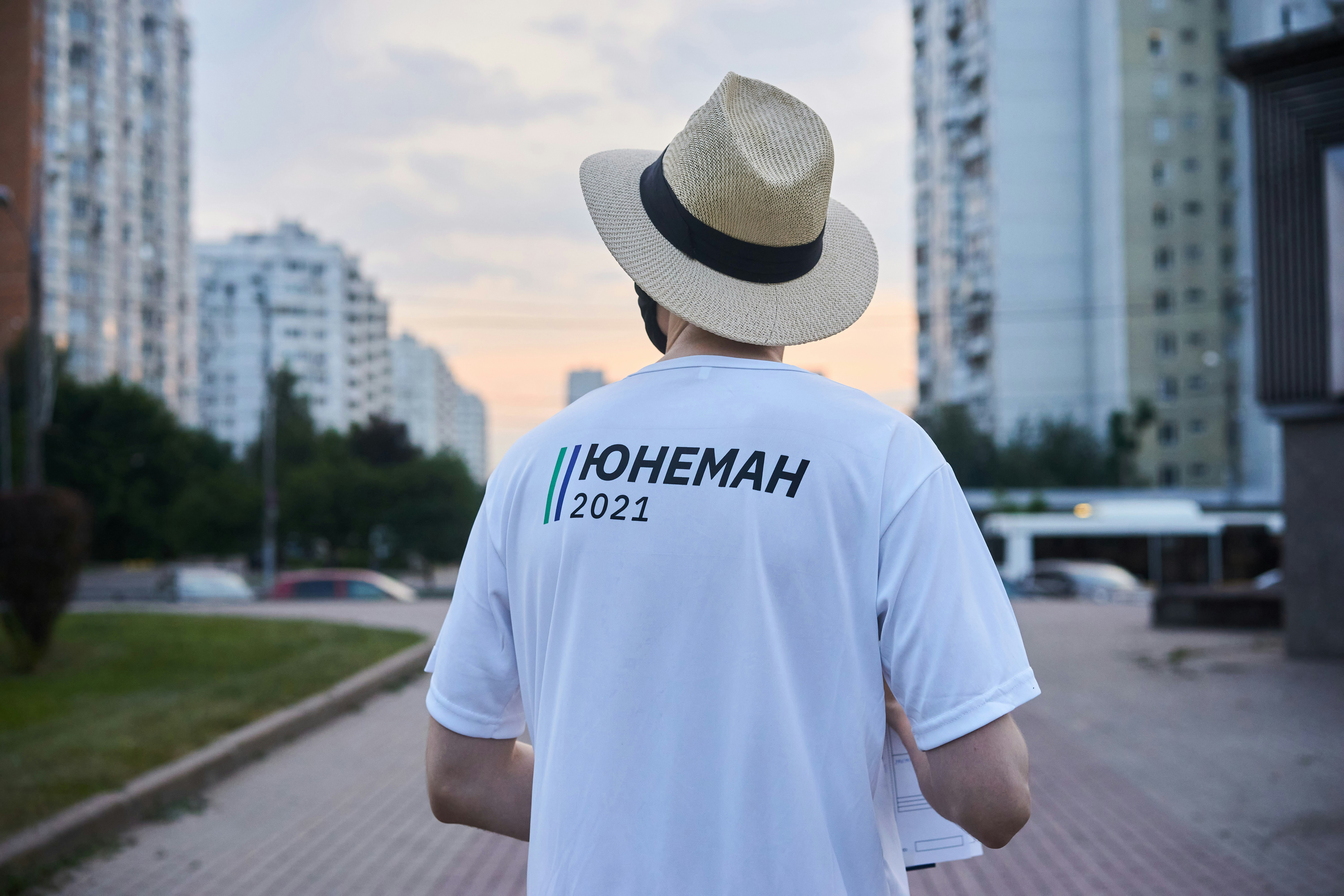 man in white crew neck t-shirt wearing brown fedora hat standing on gray concrete pavement