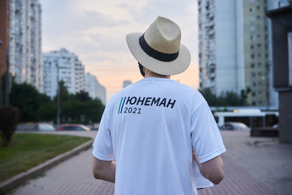 man in white crew neck t-shirt wearing brown fedora hat standing on gray concrete pavement