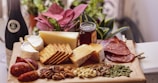 A charcuterie board features an assortment of cheeses, including a wheel of brie and blocks of hard cheese, alongside slices of cured meats and a variety of crackers. Nuts such as pecans, almonds, and cashews are scattered in front of the cheeses. A jar of honey is centrally placed, next to leafy green plants and a bottle of wine, adding a touch of elegance and nature to the setting.