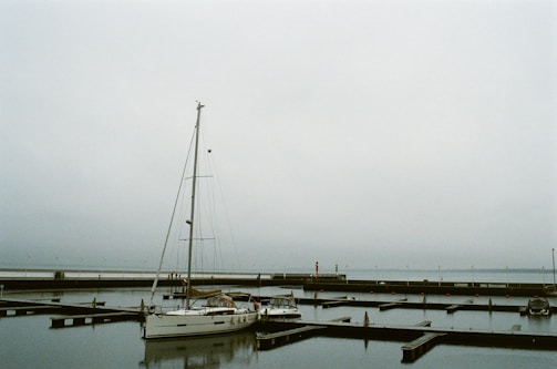 A marina with a sailboat docked in calm waters under an overcast sky. The background features a long pier and several buoys floating in the water.