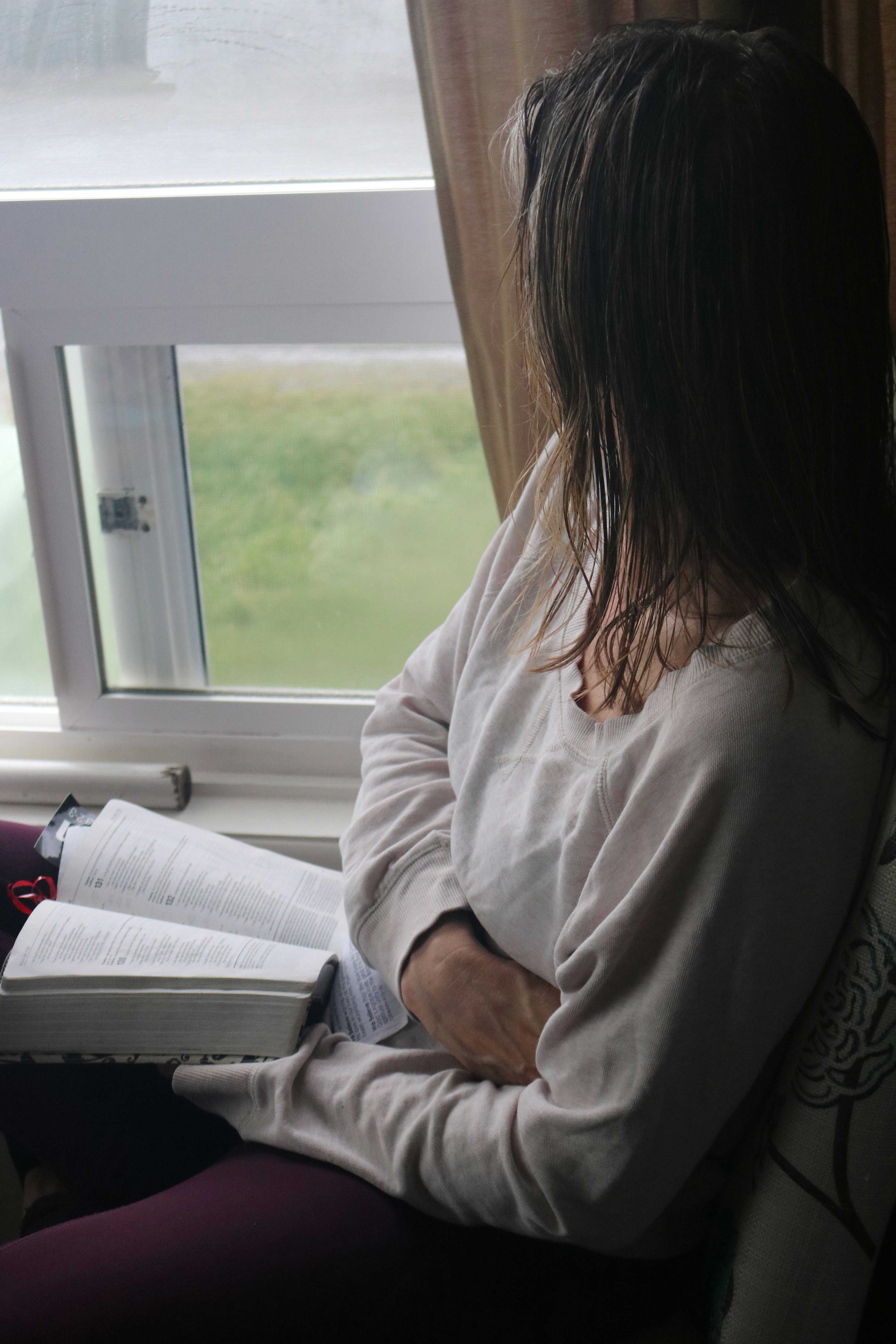 A woman sits on a window sill, reading a book, with sunlight illuminating her and the surrounding room.