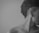 A monochrome profile of a person with short, curly hair and an undercut. The individual is wearing a long, thin earring and the image is framed to only include part of their head, ear, and shoulder against a plain background.