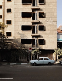 A vintage car is parked on a street in front of a building with a unique architectural design featuring rectangular protruding elements. The building's fa&ccedil;ade is made of gray stone and has multiple windows. Shadows from the building and nearby trees create an interesting play of light and dark on the surfaces.