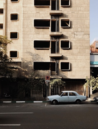 A vintage car is parked on a street in front of a building with a unique architectural design featuring rectangular protruding elements. The building's fa&ccedil;ade is made of gray stone and has multiple windows. Shadows from the building and nearby trees create an interesting play of light and dark on the surfaces.