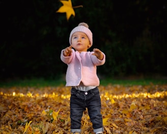 child in white sweater and blue denim jeans standing on dried leaves during daytime