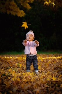 child in white sweater and blue denim jeans standing on dried leaves during daytime