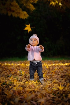 child in white sweater and blue denim jeans standing on dried leaves during daytime