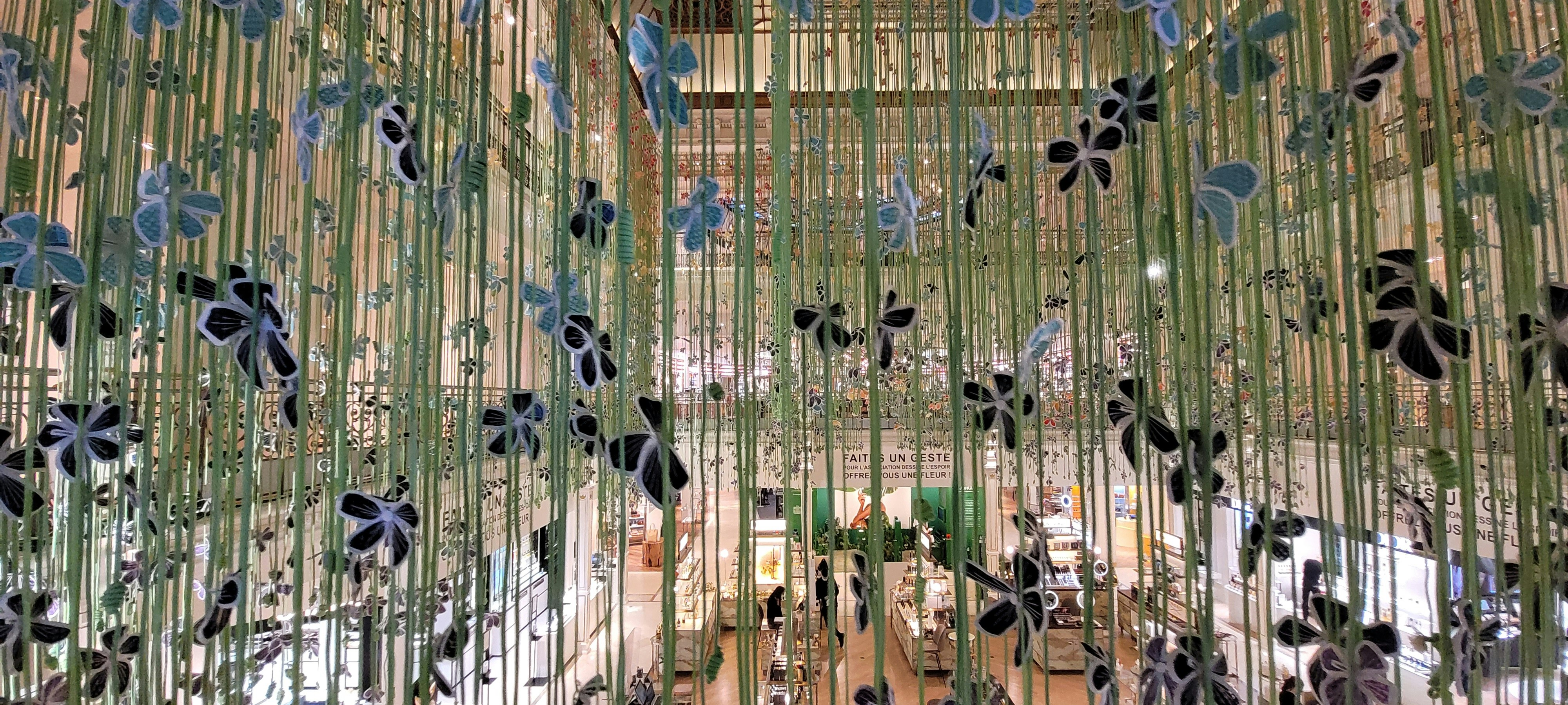 A decorative installation with vertical green strands adorned with black and blue floral-like ornaments is displayed indoors. The background shows an interior space with a high ceiling featuring a busy commercial area with various shops and people milling around.
