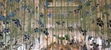 A decorative installation with vertical green strands adorned with black and blue floral-like ornaments is displayed indoors. The background shows an interior space with a high ceiling featuring a busy commercial area with various shops and people milling around.