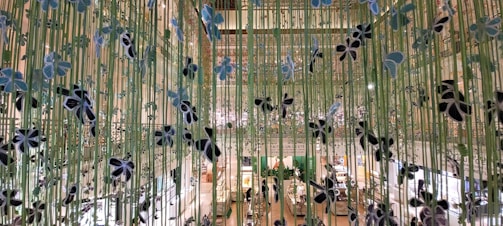 A decorative installation with vertical green strands adorned with black and blue floral-like ornaments is displayed indoors. The background shows an interior space with a high ceiling featuring a busy commercial area with various shops and people milling around.