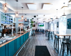 A modern bar or pub interior with high ceilings and large windows allowing natural light to flood the space. The room features high tables and chairs, with a long bar counter on the left lined with taps. The decor includes hanging pendant lights and a menu board displaying drink options. A television screen is mounted on the wall showing a sports game.