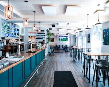 A modern bar or pub interior with high ceilings and large windows allowing natural light to flood the space. The room features high tables and chairs, with a long bar counter on the left lined with taps. The decor includes hanging pendant lights and a menu board displaying drink options. A television screen is mounted on the wall showing a sports game.