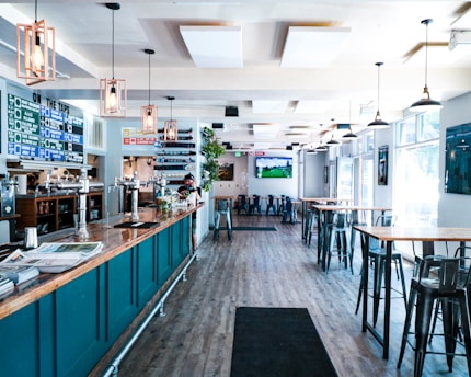 A modern bar or pub interior with high ceilings and large windows allowing natural light to flood the space. The room features high tables and chairs, with a long bar counter on the left lined with taps. The decor includes hanging pendant lights and a menu board displaying drink options. A television screen is mounted on the wall showing a sports game.