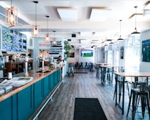A modern bar or pub interior with high ceilings and large windows allowing natural light to flood the space. The room features high tables and chairs, with a long bar counter on the left lined with taps. The decor includes hanging pendant lights and a menu board displaying drink options. A television screen is mounted on the wall showing a sports game.