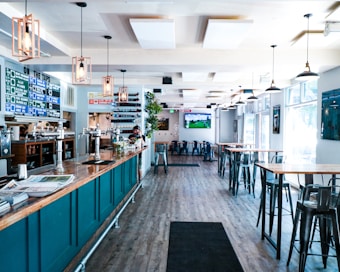 A modern bar or pub interior with high ceilings and large windows allowing natural light to flood the space. The room features high tables and chairs, with a long bar counter on the left lined with taps. The decor includes hanging pendant lights and a menu board displaying drink options. A television screen is mounted on the wall showing a sports game.