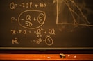 A blackboard with mathematical equations written in chalk, including variables like Q and P, and calculations involving these variables. An eraser and pieces of chalk are on the chalkboard's tray.