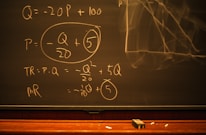 A blackboard with mathematical equations written in chalk, including variables like Q and P, and calculations involving these variables. An eraser and pieces of chalk are on the chalkboard's tray.