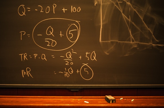 A blackboard with mathematical equations written in chalk, including variables like Q and P, and calculations involving these variables. An eraser and pieces of chalk are on the chalkboard's tray.