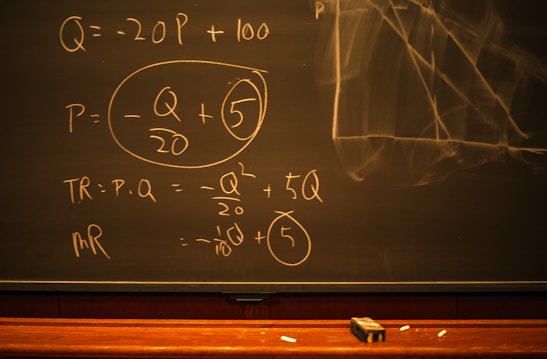 A blackboard with mathematical equations written in chalk, including variables like Q and P, and calculations involving these variables. An eraser and pieces of chalk are on the chalkboard's tray.