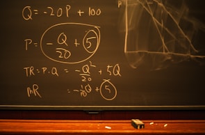 A blackboard with mathematical equations written in chalk, including variables like Q and P, and calculations involving these variables. An eraser and pieces of chalk are on the chalkboard's tray.
