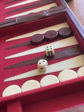 A backgammon board with red and white triangular patterns on a red background. Dark and light-colored checkers are placed on different points, and two dice are visible on the board indicating a potential move in progress.