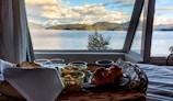 A cozy breakfast tableau is set beside a large window, revealing a stunning view of a lake with snow-capped mountains and lush forests in the background. The breakfast includes croissants, toast, and a bowl of fruit, neatly arranged in a wooden tray. Glasses and a folded napkin add to the tidy presentation.