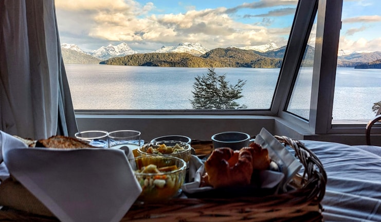 A cozy breakfast tableau is set beside a large window, revealing a stunning view of a lake with snow-capped mountains and lush forests in the background. The breakfast includes croissants, toast, and a bowl of fruit, neatly arranged in a wooden tray. Glasses and a folded napkin add to the tidy presentation.