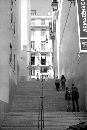 An urban scene featuring a flight of stone steps flanked by tall buildings. Several pedestrians are walking, ascending, and descending the stairs. A signage board with text in Portuguese is visible on the right side. The buildings have multiple stories with many windows and balconies.