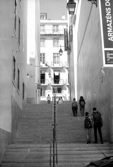 An urban scene featuring a flight of stone steps flanked by tall buildings. Several pedestrians are walking, ascending, and descending the stairs. A signage board with text in Portuguese is visible on the right side. The buildings have multiple stories with many windows and balconies.