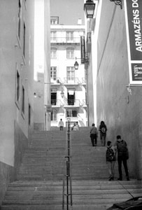 An urban scene featuring a flight of stone steps flanked by tall buildings. Several pedestrians are walking, ascending, and descending the stairs. A signage board with text in Portuguese is visible on the right side. The buildings have multiple stories with many windows and balconies.