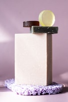 A set of soap bars arranged in a minimalistic style. A large rectangular off-white soap stands vertically, topped with smaller round soaps in green, brown, and yellow hues. The soaps rest on a textured lavender mat, against a softly lit lavender background.